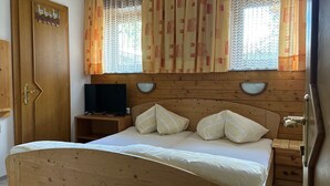 A wooden bed with a headboard, two bedside tables, a television on a stand, and a window with patterned curtains. - Studio apartment - House Luxer (Auffach)
