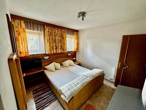 A single bed with a wooden headboard, a television on a stand, and a window with patterned curtains. - Studio apartment - House Luxer (Auffach)