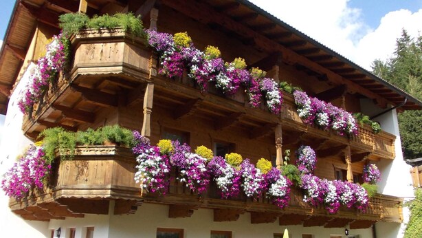A wooden building with balconies adorned with colorful flowers and a covered outdoor seating area. - Studio apartment - House Luxer (Auffach)