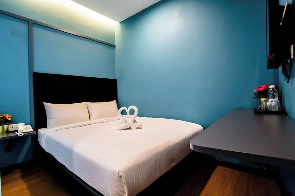 A hotel room with a bed, a bedside table with a phone, a small table with a bottle and flowers, and a wall-mounted TV.