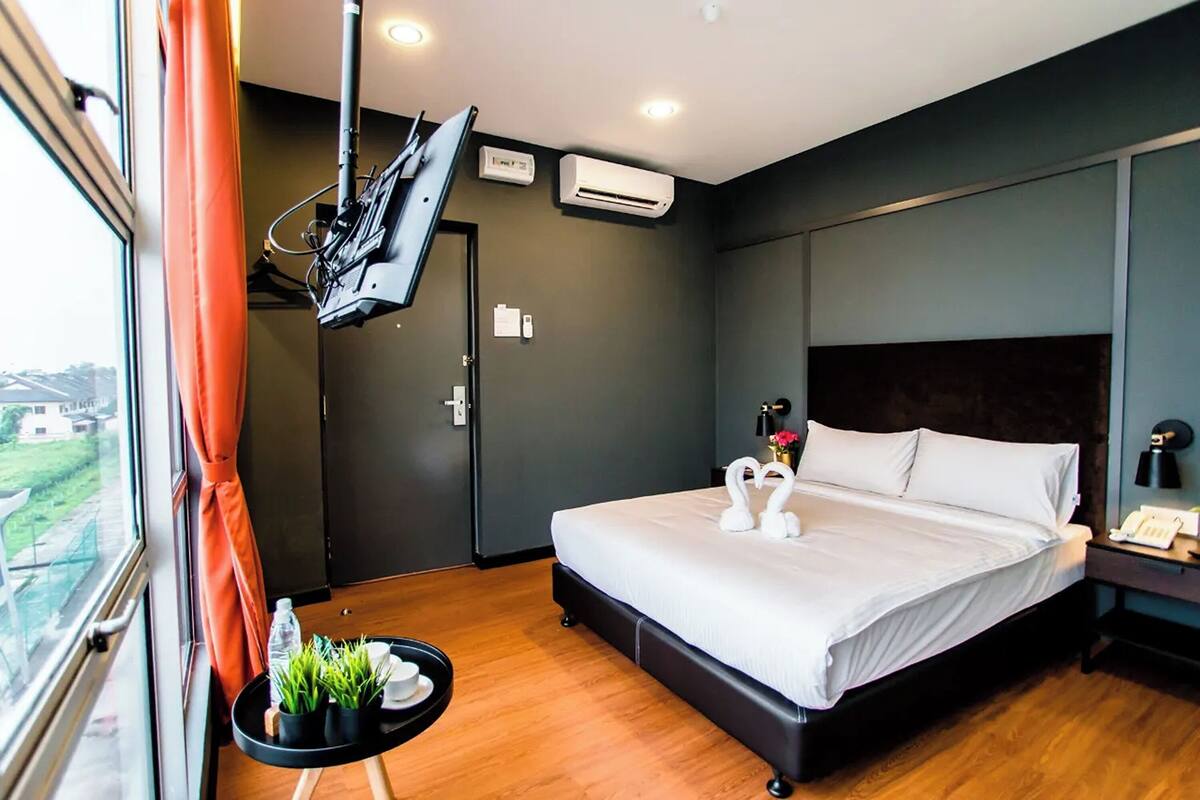 A hotel room with a bed, a TV mounted on the wall, an air conditioning unit, and a window with curtains.