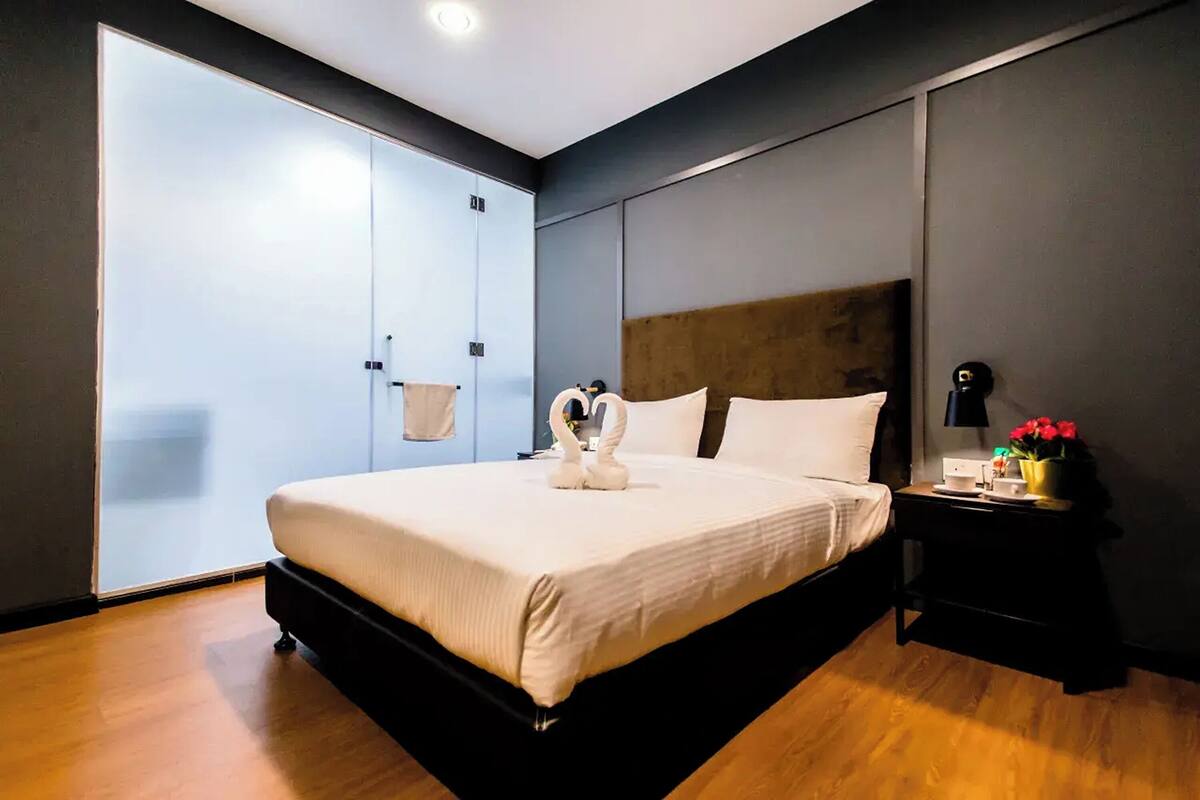 A modern bedroom with a large bed, a wooden floor, a sliding door, and a nightstand with a lamp and flowers.