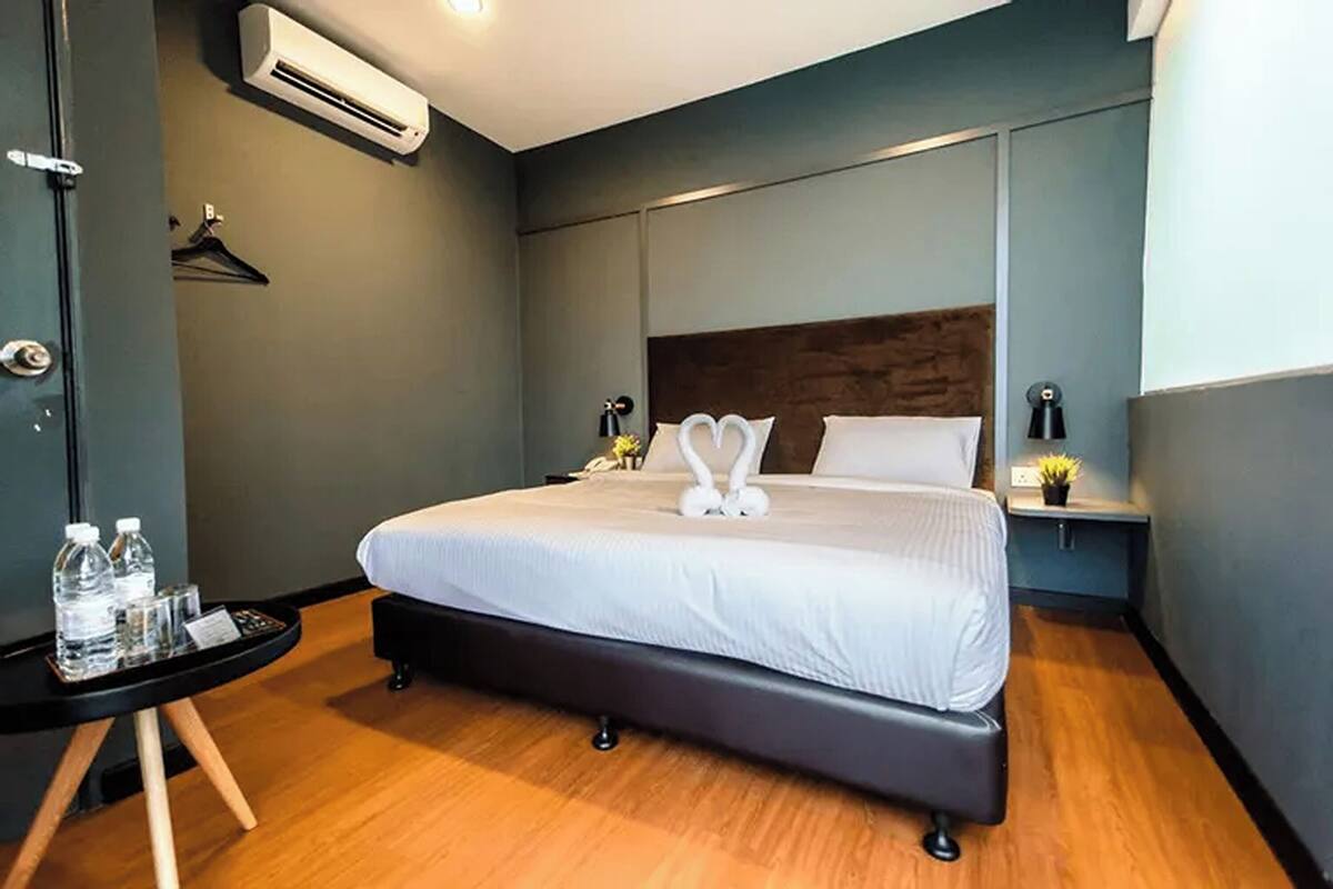 A modern hotel room with a large bed, a bedside table with a lamp, and a small table with water bottles and a menu.