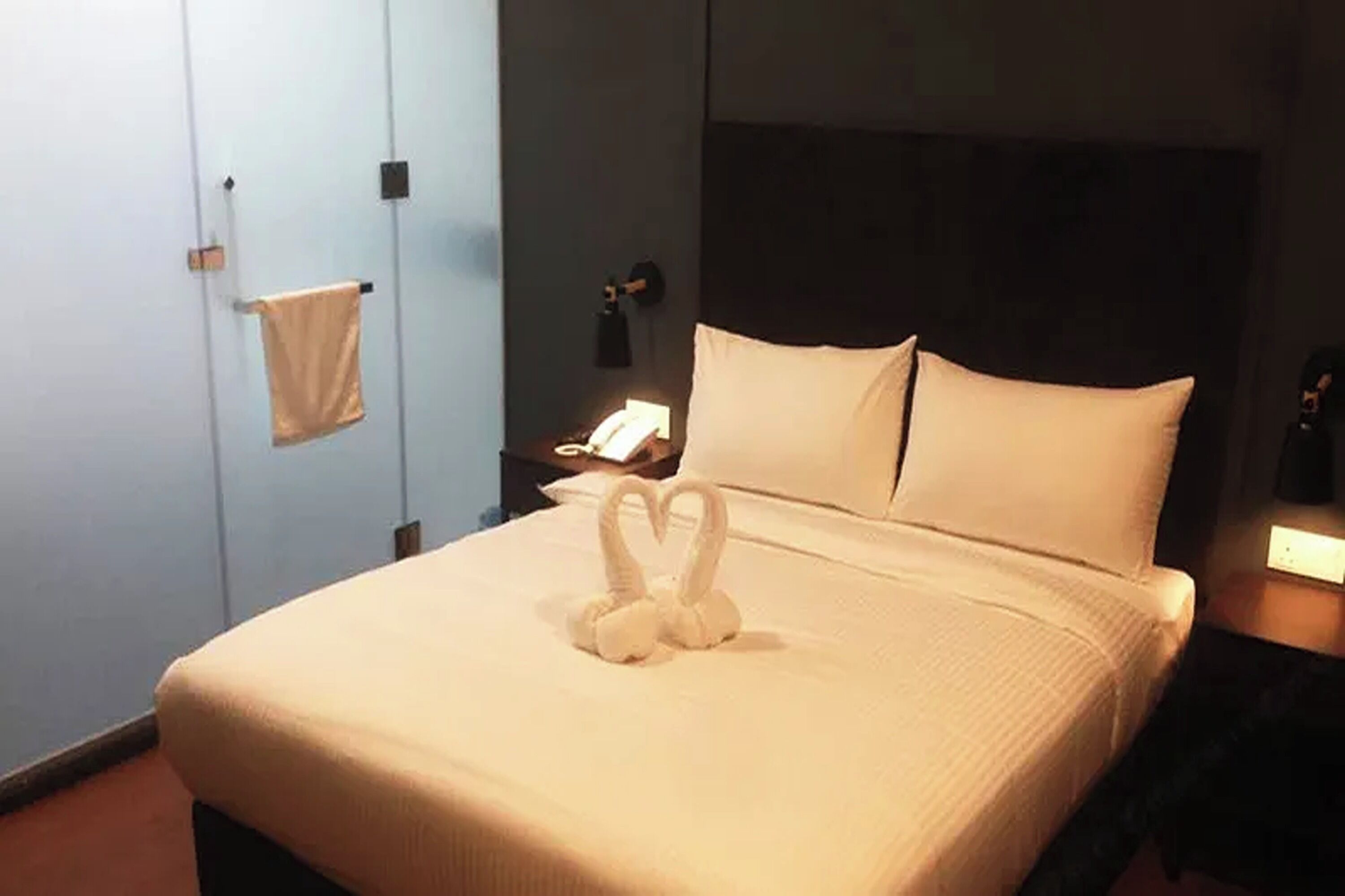 Una cama bien hecha con una decoración de toalla con forma de corazón, una mesita de noche con lámpara y un teléfono fijado a la pared.