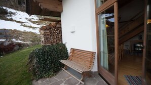 A wooden bench outside a building with stone flooring, a white wall, and a glass door leading inside.