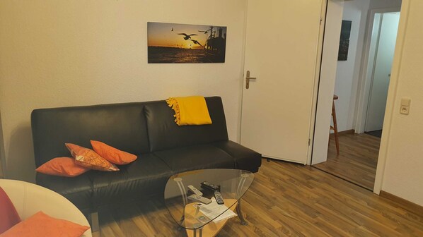 A living room with a black leather sofa, two orange pillows, a glass coffee table with remotes and a magazine, and a painting of birds flying over water on the wall.