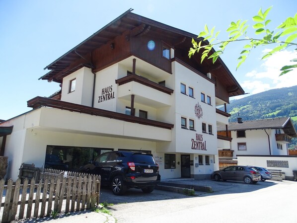 A multi-story building with 'HAUS ZENTRAL' signage, a parking area, and a mountainous backdrop.