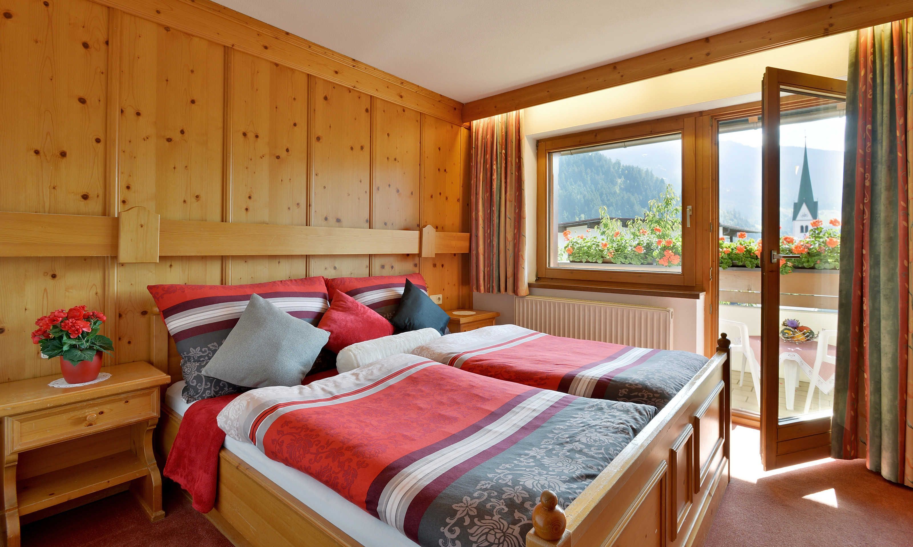 Ein Zimmer mit Holzwänden, zwei Betten, einem Nachttisch mit Pflanze und einem Fenster mit Blick auf Berge und Blumen.