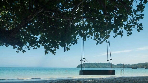 A beach with a swing set under a tree, clear blue water, and a distant island. - Payam Cottage Resort (Ranong)
