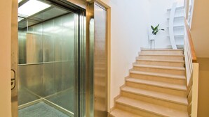 Un ascensor con pantalla digital, una escalera con planta y una pared blanca.