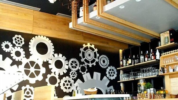 Zona de bar con mesas y taburetes de madera, barra con botellas y vasos, y pared decorada con diseños de engranajes blancos.