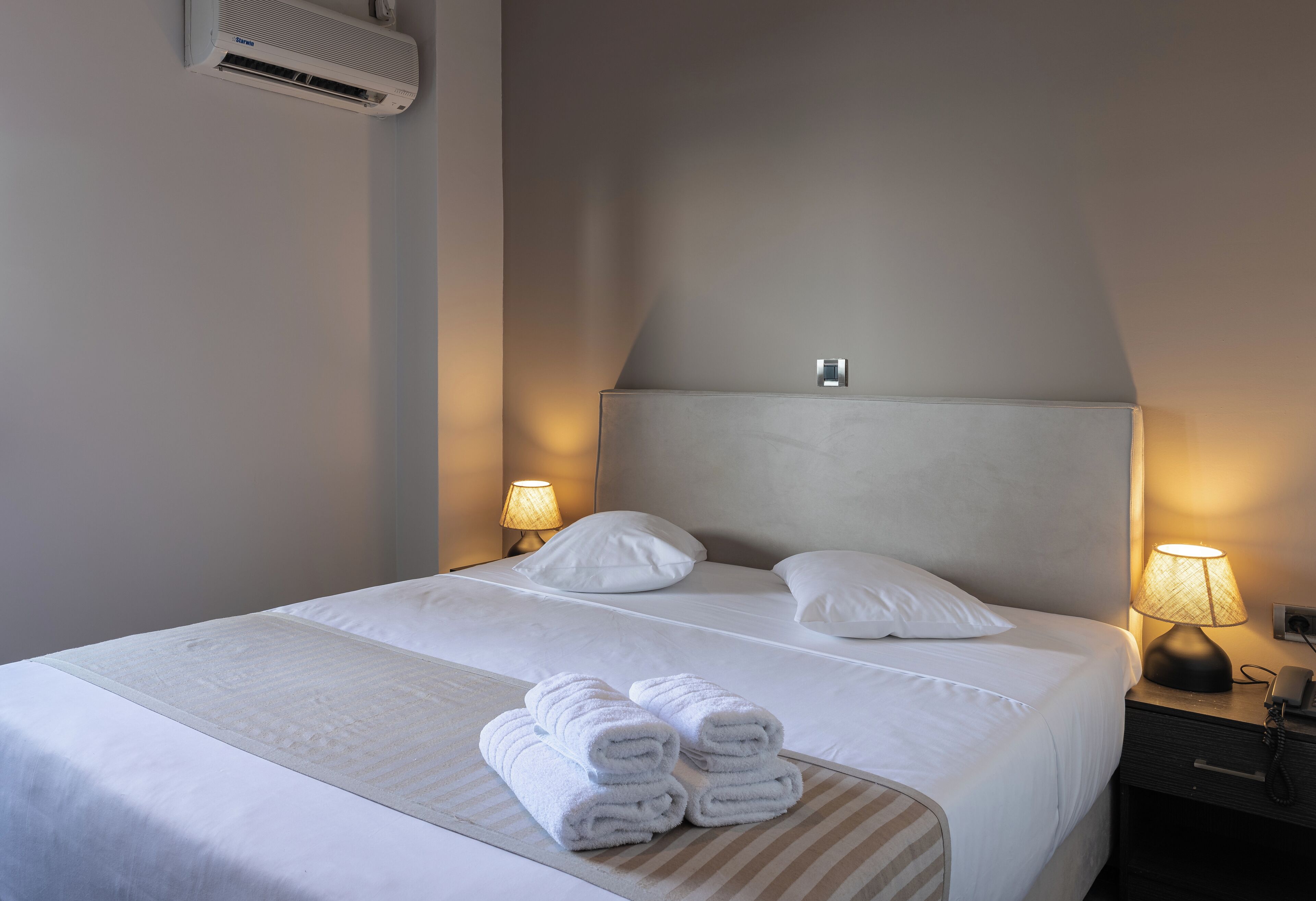 Een netjes opgemaakt bed met witte lakens en opgerolde handdoeken, een bedlamp en een airconditioningunit aan de muur.