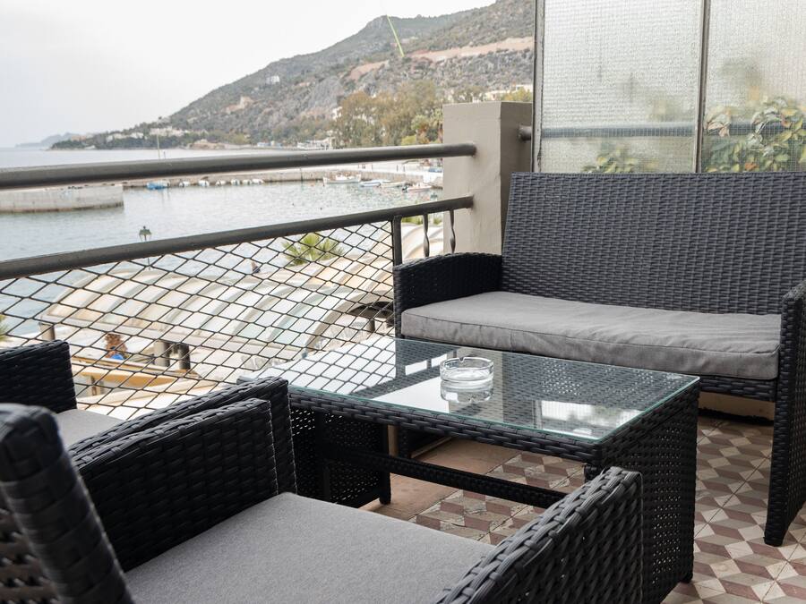A balcony with wicker furniture, a glass table, and a view of the sea and hills.