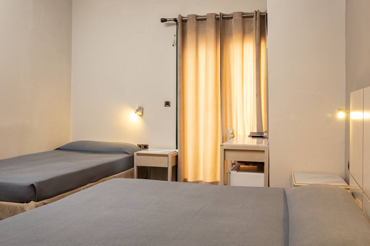 A hotel room with two single beds, a small bedside table, a nightstand, a mirror, and a curtain.