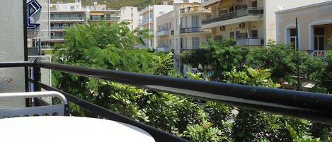 A balcony with a white table, surrounded by greenery and overlooking a row of buildings with balconies, set against a mountainous backdrop.