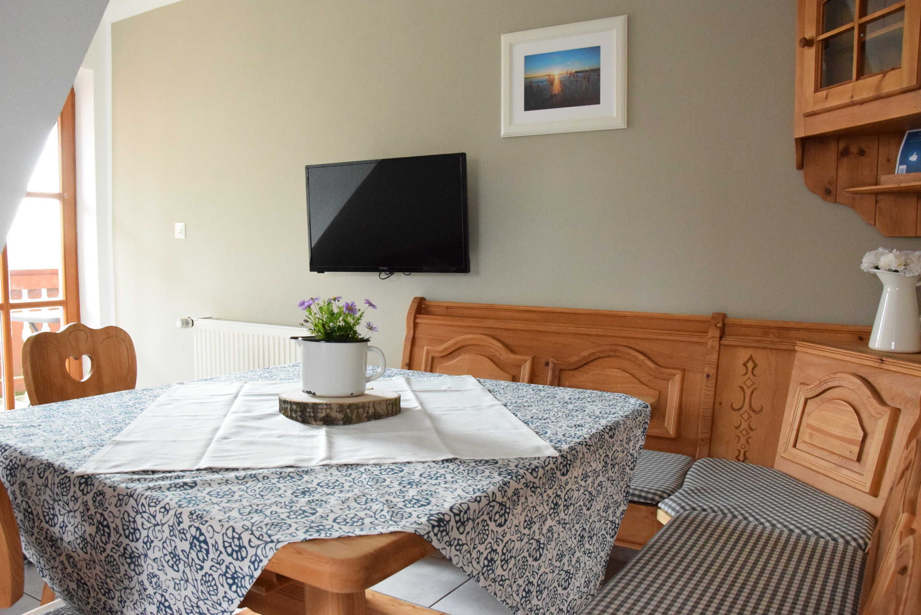 A room with a bed, a table with a patterned tablecloth, a television mounted on the wall, and a framed picture above the television.