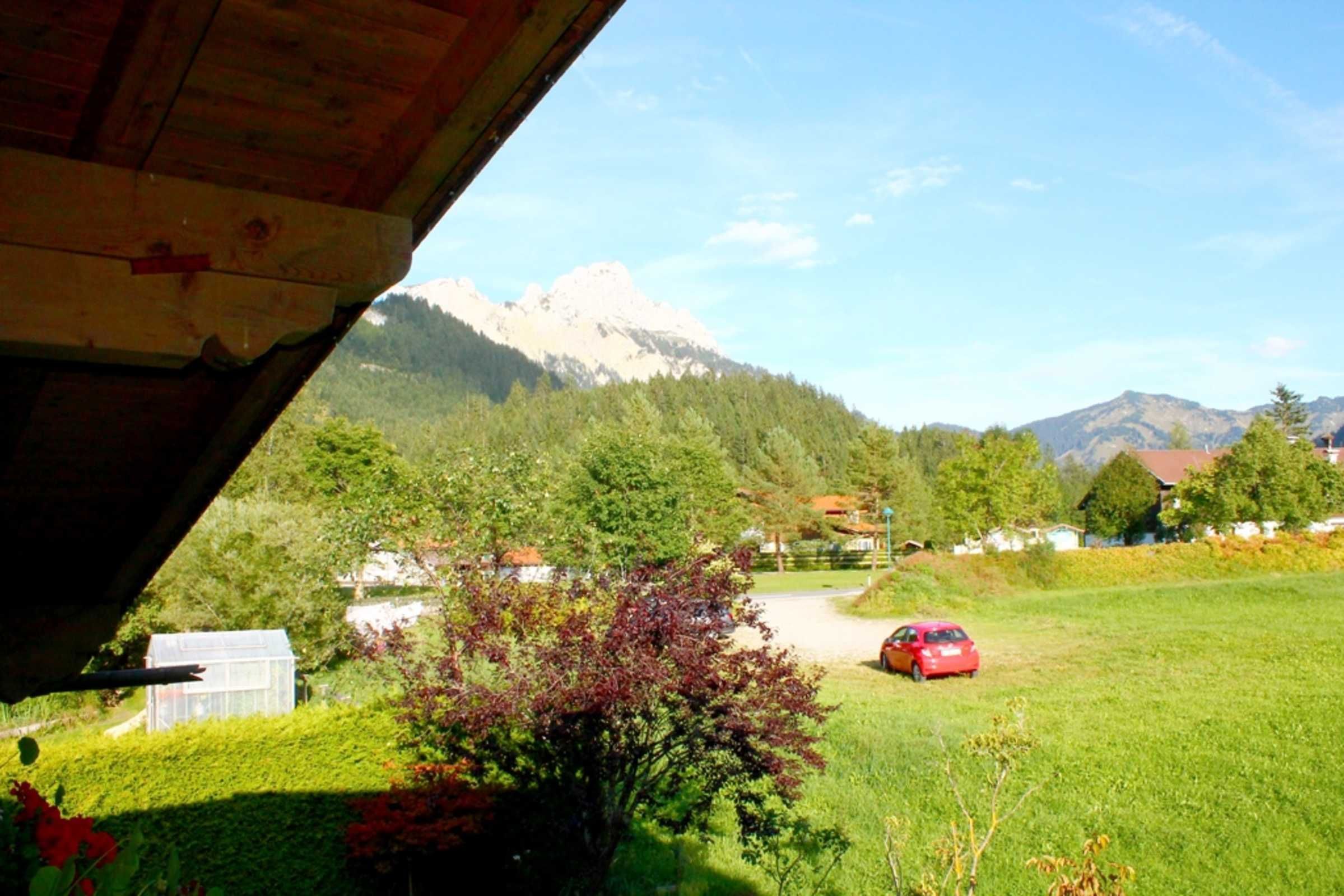 Blick auf eine gebirgige Landschaft, mit einem roten Auto auf einer Wiese, einem Gewächshaus und einem Gebäude mit geneigtem Dach.