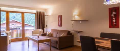 Una sala de estar moderna con sofá, mesa de centro, mesa de comedor, sillas, una lámpara colgante y vista a un edificio a través de amplios ventanales.