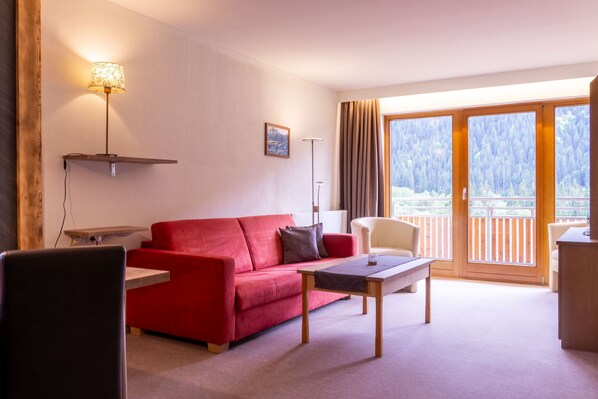 A living room with a red sofa, a wooden coffee table, a lamp, and a view of a forest through large windows.
