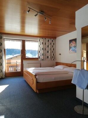 A bedroom with a wooden bed, a window with curtains, a radiator, and a painting on the wall. - Künzelspitze DZ#2 - Ritter Anni - Haus Ritter (Schoppernau)