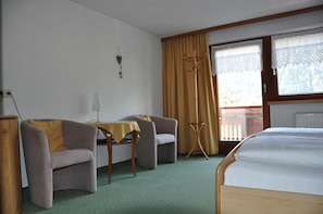 A hotel room with a bed, two armchairs, a small table, a coat rack, and a window with curtains.