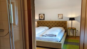 Habitación de hotel con cabina de ducha de vidrio, cama con ropa blanca, piso de madera y una pequeña mesa con lámpara.