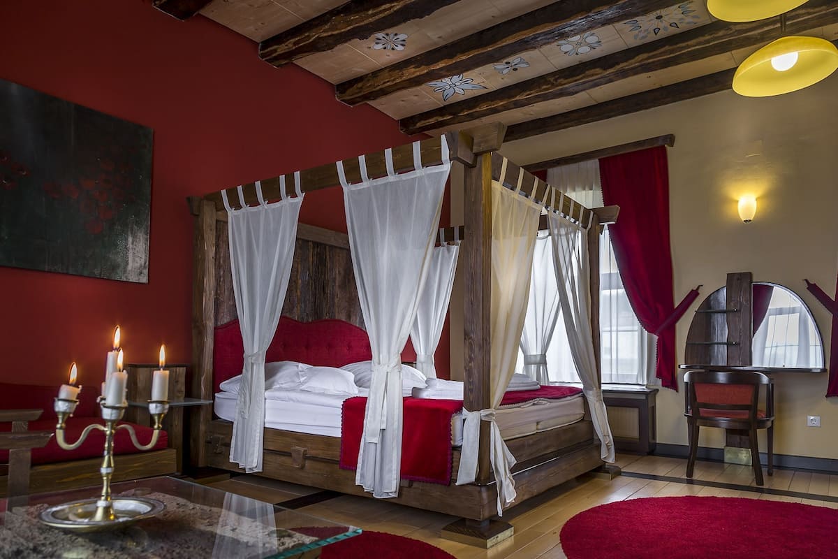 A four-poster bed with white drapes, a wooden headboard, a red accent wall, a glass table with candles, and a red rug.