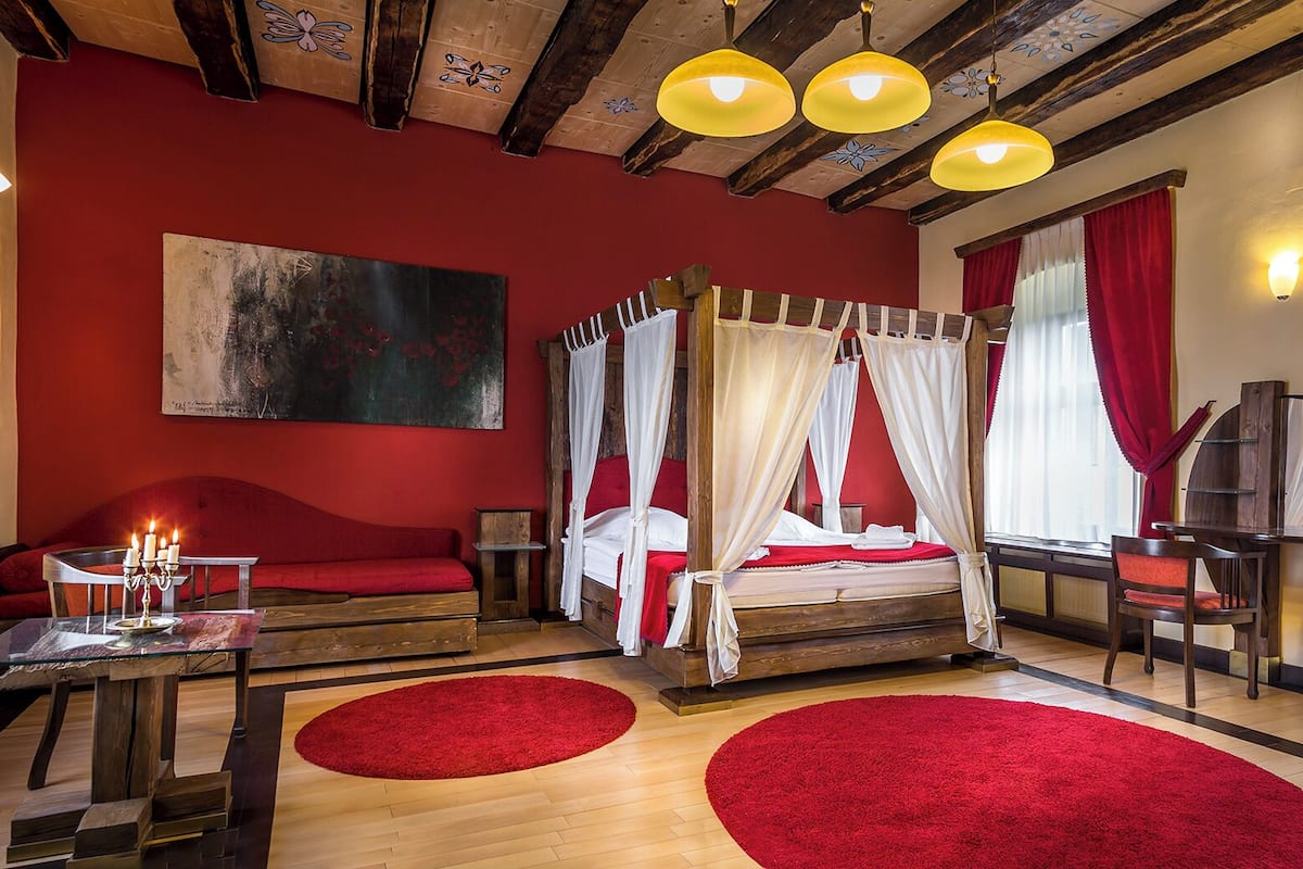A room with a four-poster bed, red walls, and wooden beams. There are two red rugs on the floor, a table with candles, and a painting on the wall.