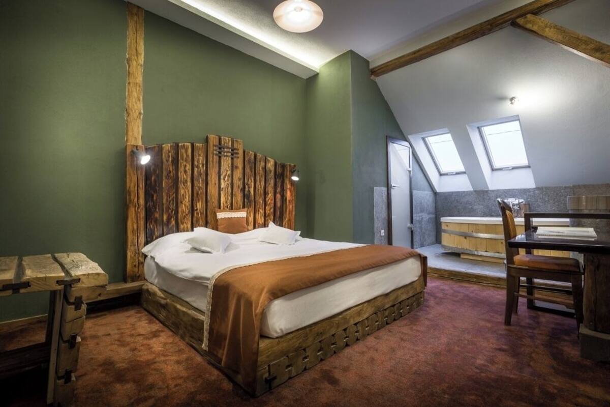A bedroom with a wooden headboard, a bed, a wooden bench, a table, and a view of a bathroom with a bathtub and skylights.