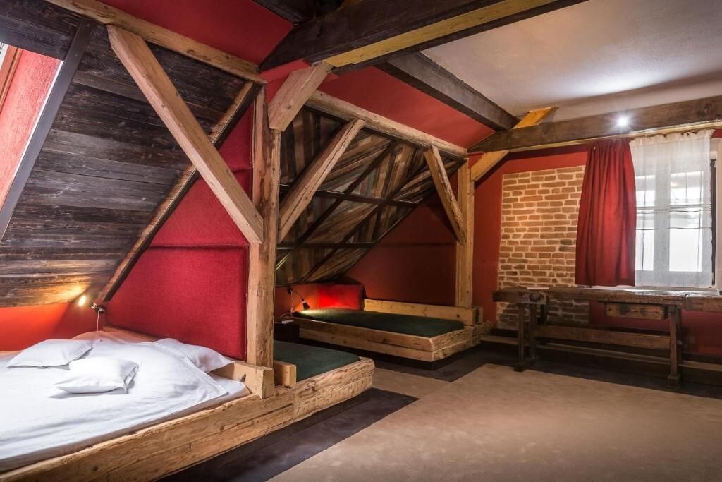 A rustic attic bedroom with exposed wooden beams, a brick wall, and a window with red curtains.