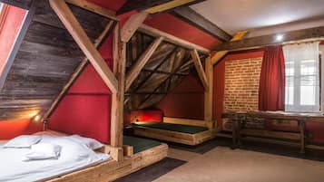 A rustic attic bedroom with exposed wooden beams, a brick wall, and a window with red curtains.