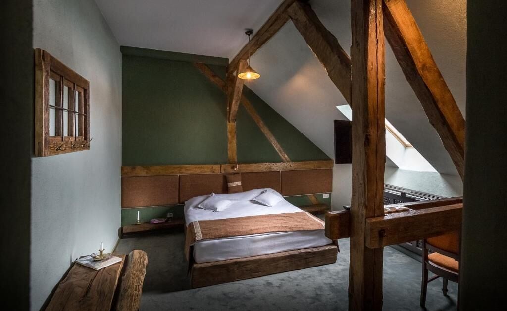 Quarto estilo loft com vigas de madeira aparentes, cama com roupa de cama branca, pequena mesa com uma vela e vista do céu através de um claraboia.