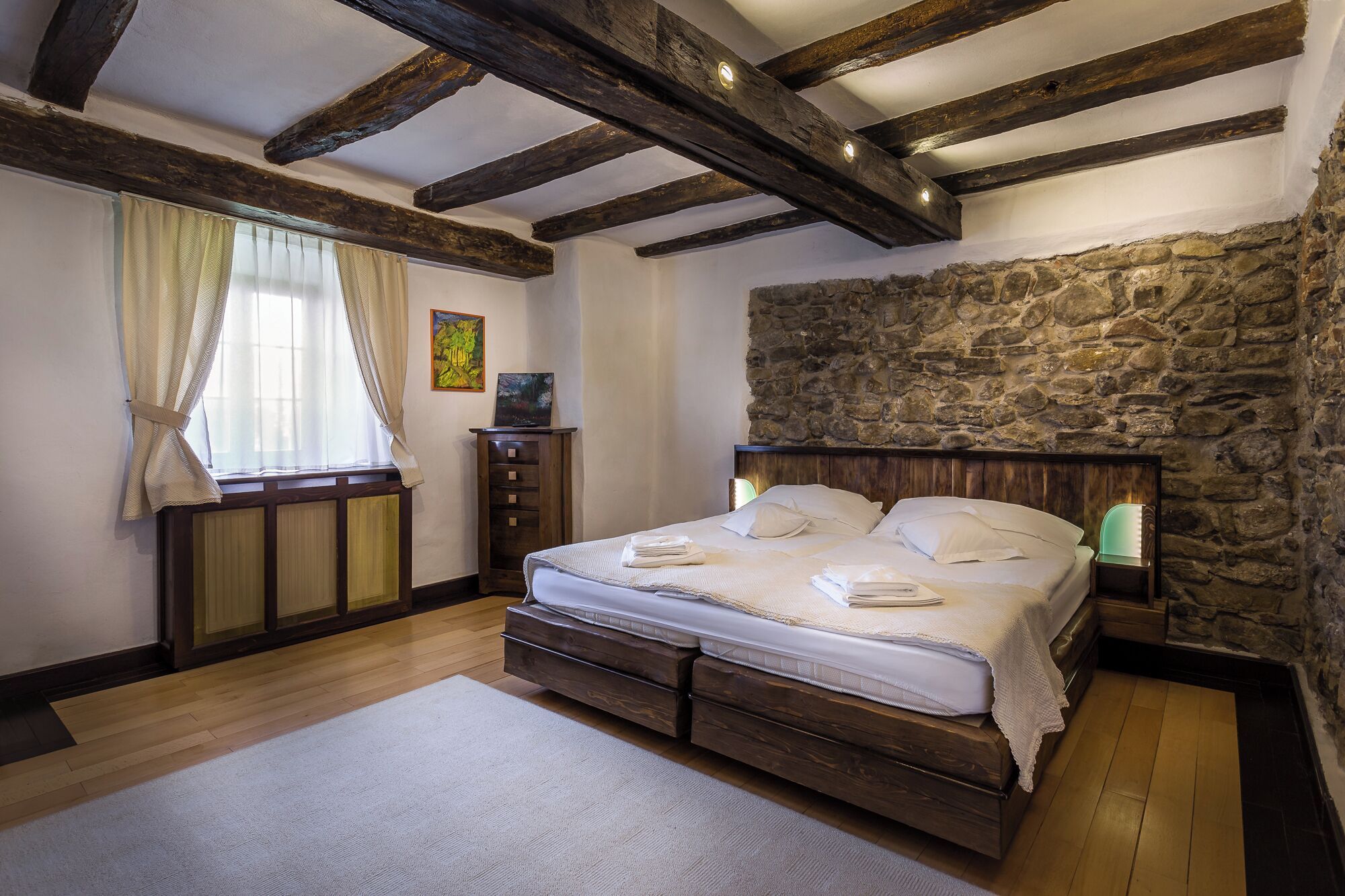 Une chambre avec un mur en pierre, des poutres apparentes, un lit avec du linge de maison blanc, une table de chevet et une fenêtre avec des rideaux.