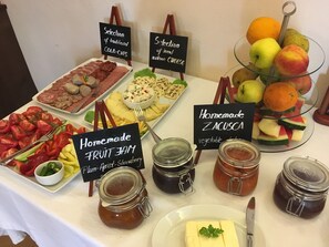 Une table de buffet proposant une variété de plats, notamment de la confiture de fruits faite maison, de la charcuterie, des fromages artisanaux et des fruits frais comme des pommes et des oranges.