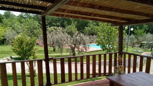 A wooden deck with a table and bench, overlooking a garden with a pool and trees. - Il Poggio (Pontedera)