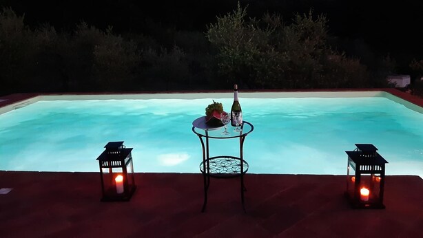 A swimming pool at dusk with two tables, each with a bottle and glass, and two lit lanterns. - Il Poggio (Pontedera)