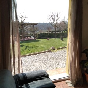 Vista desde adentro de una habitación con un ventanal grande que da a un jardín con césped, un camino de grava y una pérgola.