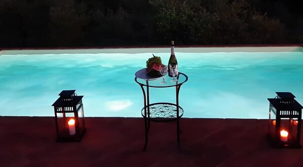 Mesa junto a la piscina con una botella y un vaso, rodeada de faroles, con vista a la piscina.