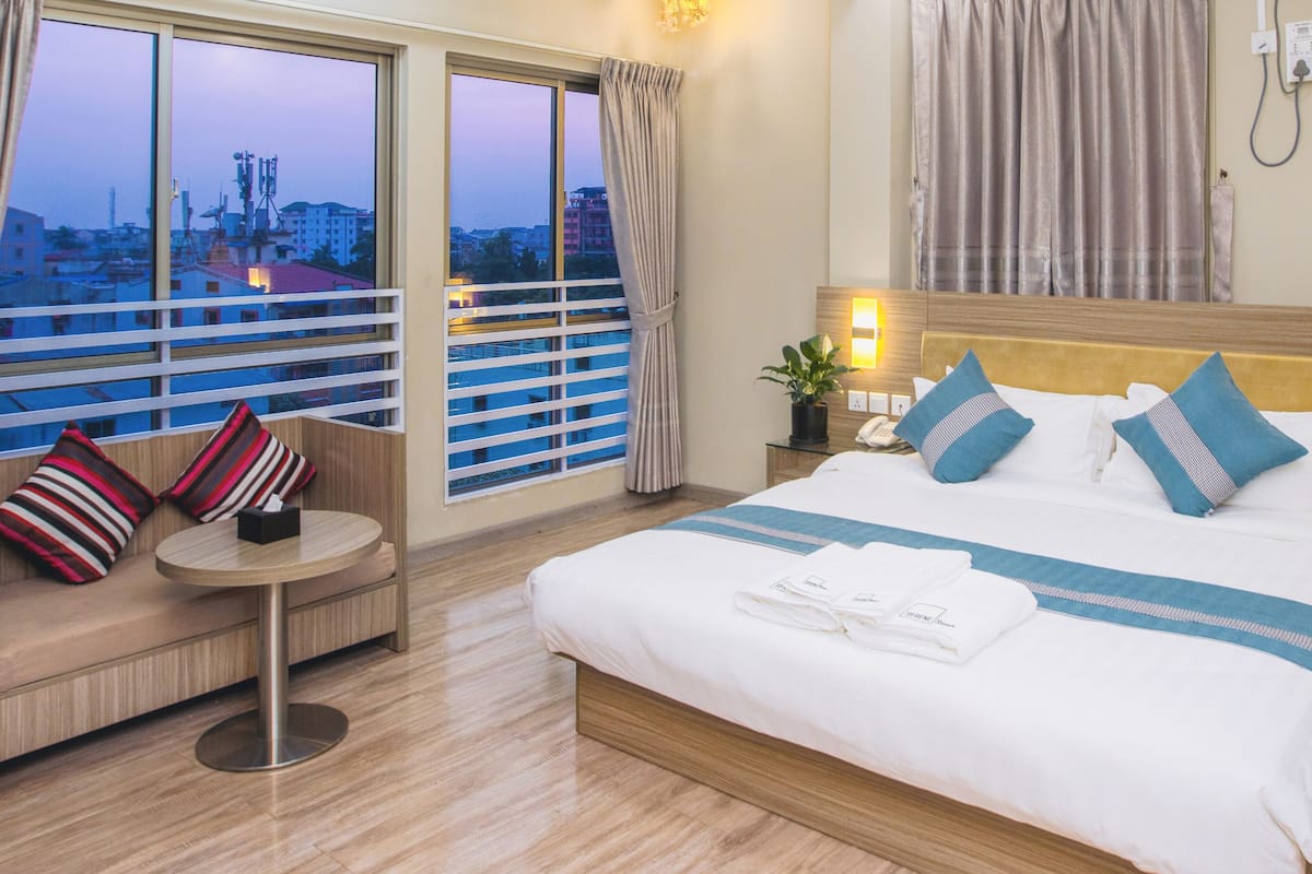 Habitación de hotel con una cama grande, un sofá, una mesita y vistas a la ciudad a través de las ventanas.