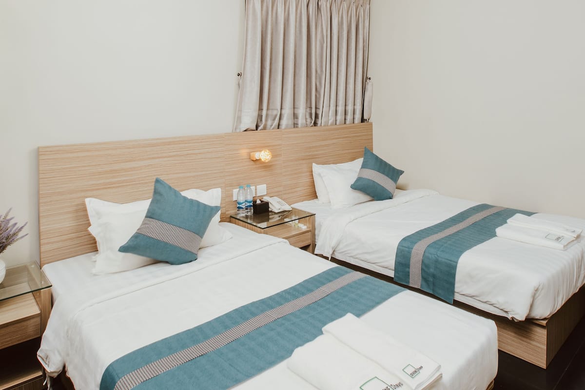 Habitación de hotel con dos camas, una mesita de noche con lámpara, una pequeña mesa con planta y un refrigerador.