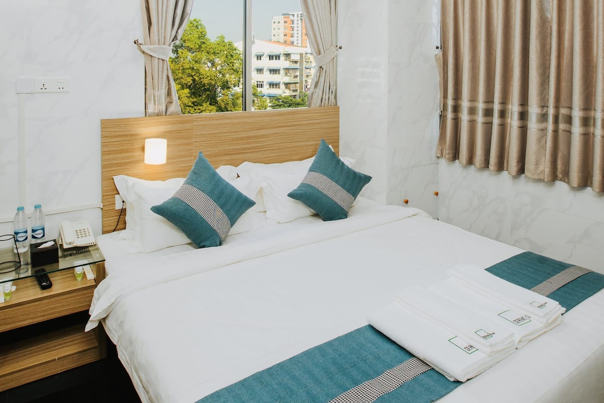 Habitación de hotel con una cama grande, cabecera de madera, mesita de noche con un teléfono y botellas de agua, y vistas a edificios a través de la ventana.