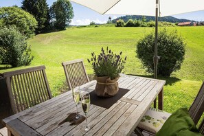 Ein Holztisch im Freien mit zwei Weingläsern, einem Topf Lavendel und Blick auf ein grünes Feld sowie entfernte Gebäude.