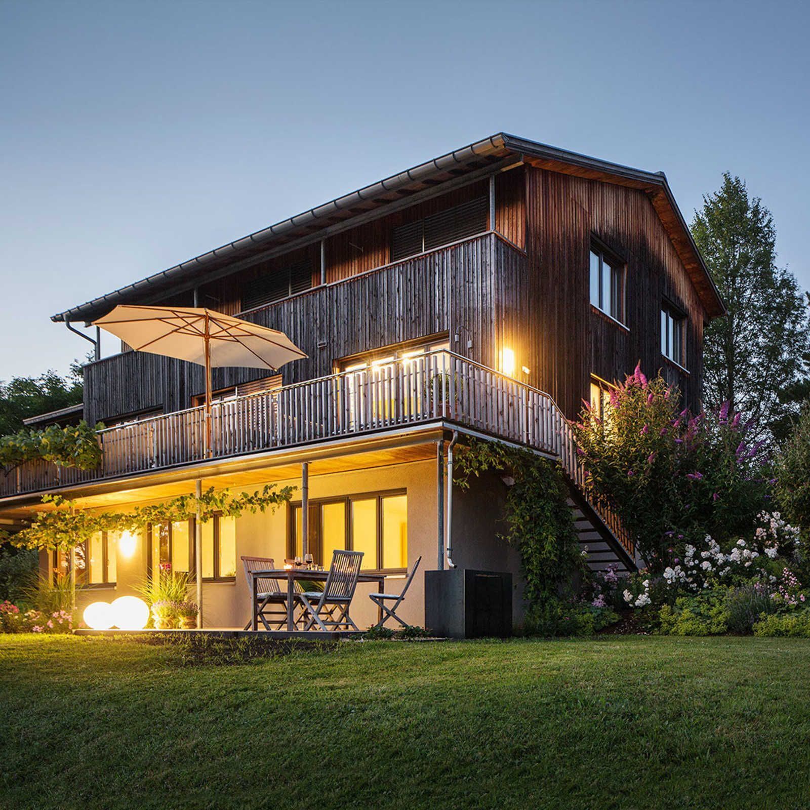 Une maison en bois à deux étages, dotée d’un balcon, d’un espace extérieur aménagé pour s’asseoir et d’un jardin bien éclairé.