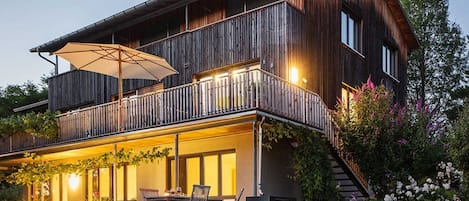 A two-story wooden house with a balcony, outdoor seating, and a well-lit garden area.