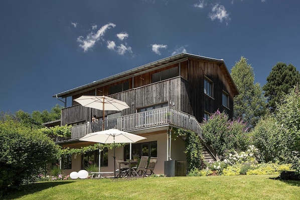 A two-story wooden house with a balcony, outdoor seating, and a garden.