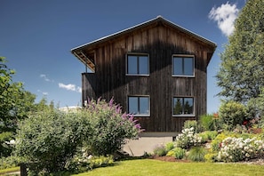 A modern two-story house with a wooden exterior, large windows, and a well-maintained garden with various plants and flowers.