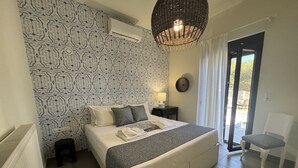 A bedroom with a patterned wallpaper, a bed with pillows, a bedside table with a lamp, a chair, and a view through a door. - Villa Ikaros (Rethymno)