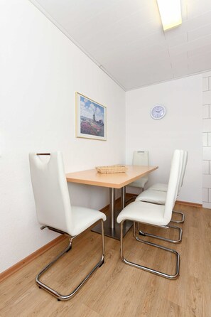Une pièce avec une table en bois, deux chaises blanches aux pieds chromés, une horloge au mur et un tableau encadré au-dessus de la table.