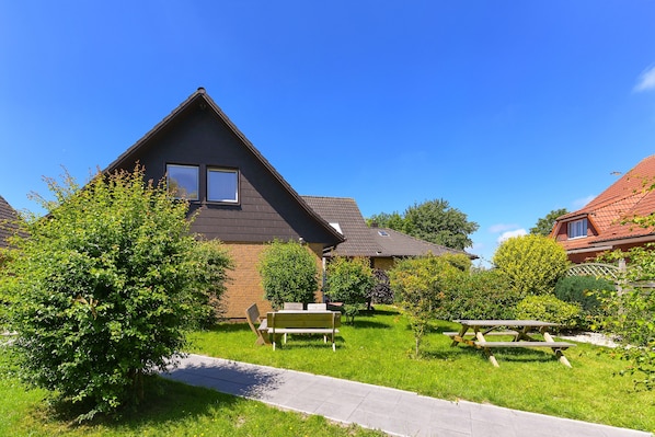 Une maison d’habitation avec un toit en pente, une cour avant avec une table de pique-nique et des bancs, et un ciel bleu dégagé.