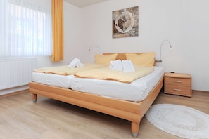 Un lit double avec une literie blanche et jaune, une table de chevet en bois avec des tiroirs, un tapis blanc et un tableau accroché au mur.